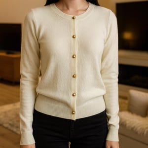 以下が含まれることがあります： 丸いネックラインと長袖のクリーム色のカーディガン。セーターは前面に金色のボタンが並んでいます。裾と袖口はリブ編みです。黒いパンツを履いています。