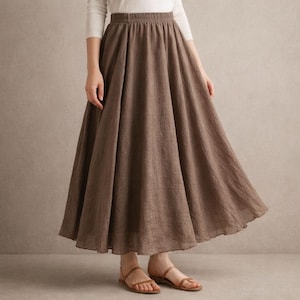 May include: A long, flowing brown skirt with an elastic waistband. The skirt is made of a textured fabric and has a wide, flared silhouette. The model is wearing brown sandals and a white long-sleeve shirt.