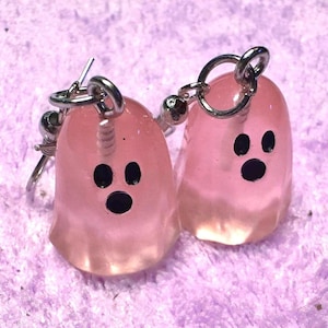 Puede incluir: Un par de pendientes de fantasma rosas con ojos y bocas negras. Los pendientes están hechos de un material translúcido y tienen aros de metal plateado. Los pendientes están sobre una superficie morada.