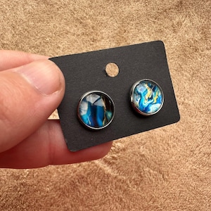May include: A pair of round stud earrings with abstract blue, white, and yellow designs set in silver-tone metal. The earrings are displayed on a black card with a small hole.