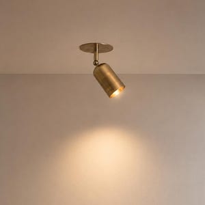 May include: A cylindrical brass spotlight fixture mounted to the ceiling. The light features a circular base plate, a short vertical stem, and an adjustable head that emits a warm, focused beam of light downward onto the wall surface below.