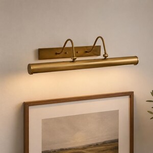 May include: A wall-mounted picture light with a brushed brass finish, featuring a horizontal cylindrical lamp head attached to a rectangular backplate by two curved arms. The fixture emits a warm, downward-facing glow over a framed artwork.
