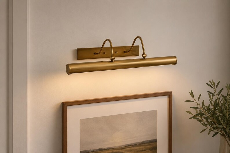 May include: A wall-mounted picture light with a brushed brass finish, featuring a horizontal cylindrical lamp head attached to a rectangular backplate by two curved arms. The fixture emits a warm, downward-facing glow over a framed artwork.