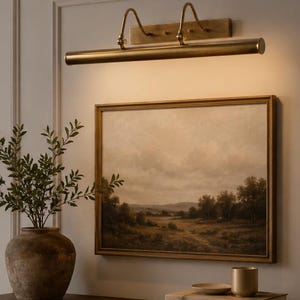 May include: A brass picture light fixture mounted above a framed landscape painting. The painting depicts a field with trees under a cloudy sky. Below, a ceramic vase, a small bowl, a metal cup, and two books titled The Kinfolk Home and Live Beautifully.