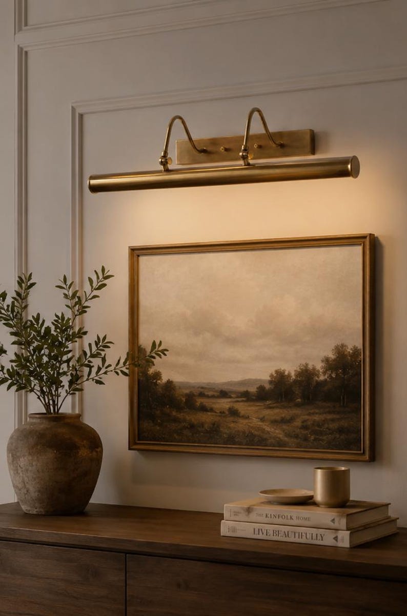 May include: A brass picture light fixture mounted above a framed landscape painting. The painting depicts a field with trees under a cloudy sky. Below, a ceramic vase, a small bowl, a metal cup, and two books titled The Kinfolk Home and Live Beautifully.