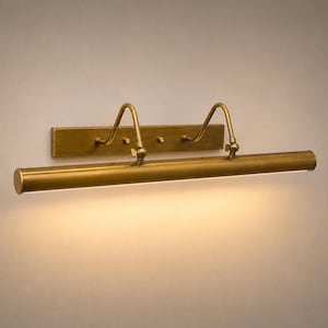 May include: A wall-mounted picture light with a brushed brass finish. The fixture features a rectangular backplate, two curved support arms with adjustable joints, and a long cylindrical lamp head that emits a warm downward glow.