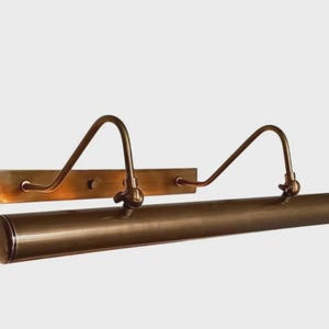 May include: A wall-mounted picture light with a long, cylindrical horizontal shade and a rectangular backplate. The fixture is finished in a brushed bronze tone and features two curved arms that connect the backplate to the light housing.