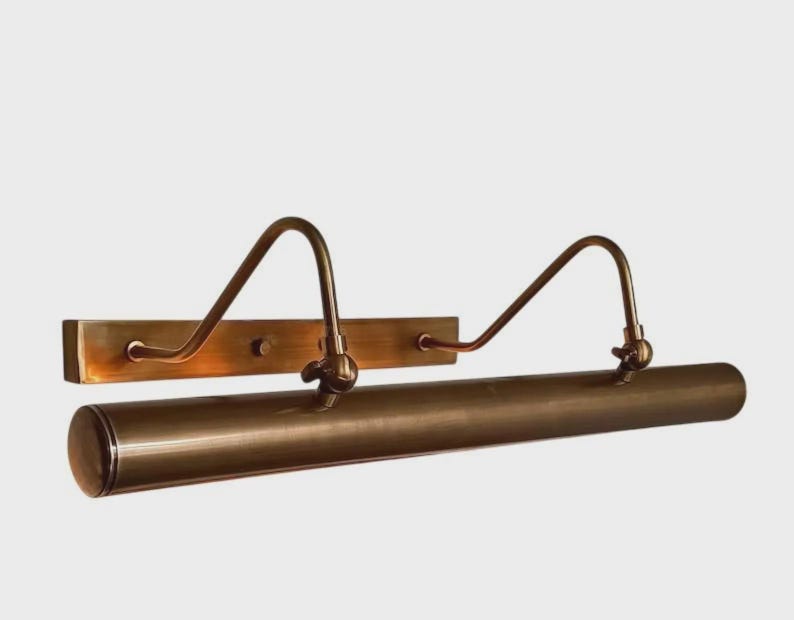 May include: A wall-mounted picture light with a long, cylindrical horizontal shade and a rectangular backplate. The fixture is finished in a brushed bronze tone and features two curved arms that connect the backplate to the light housing.