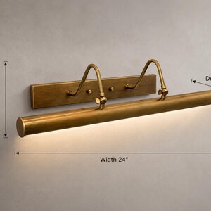 May include: A brass picture light with a rectangular wall mount, two curved support arms, and a cylindrical lamp head. The fixture measures 24 inches in width, 8 inches in height, and 5.5 inches in depth, featuring an integrated light source.
