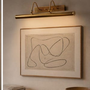 May include: A brass picture light fixture mounted above a framed rectangular artwork featuring a black continuous line drawing on a beige background. The light emits a warm glow downward, illuminating the print and the wall surface below it.