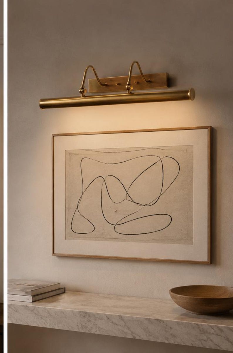 May include: A brass picture light fixture mounted above a framed rectangular artwork featuring a black continuous line drawing on a beige background. The light emits a warm glow downward, illuminating the print and the wall surface below it.