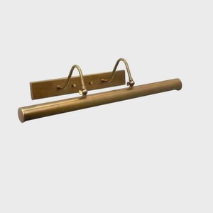 May include: A brass picture light fixture featuring a long, cylindrical horizontal tube attached to a rectangular wall-mounting plate by two curved, adjustable metal arms. The metal has a smooth, polished finish with a warm, golden-brown tone.