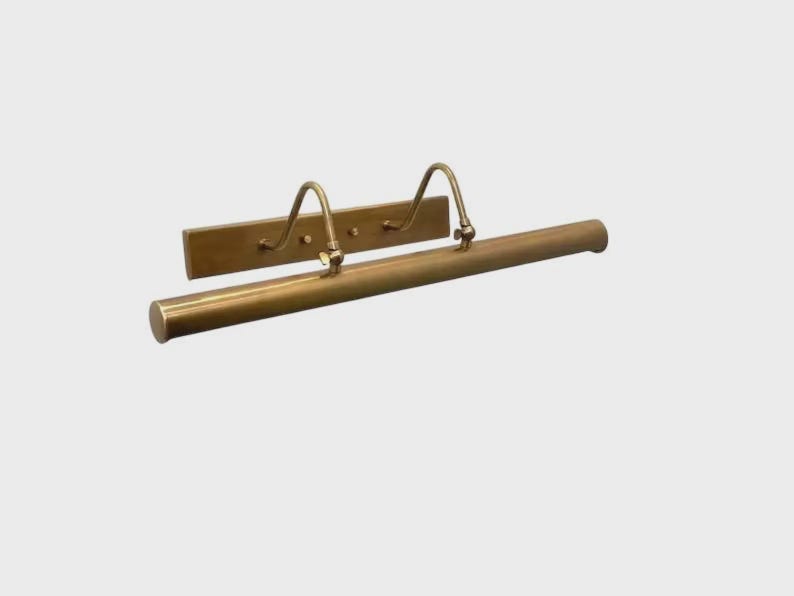 May include: A brass picture light fixture featuring a long, cylindrical horizontal tube attached to a rectangular wall-mounting plate by two curved, adjustable metal arms. The metal has a smooth, polished finish with a warm, golden-brown tone.