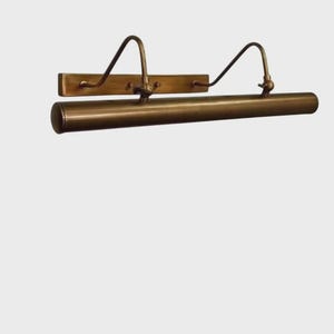May include: A horizontal picture light fixture with an antique brass finish. The design features a rectangular wall-mounting plate, two curved support arms, and a long cylindrical light housing that extends across the front of the fixture.