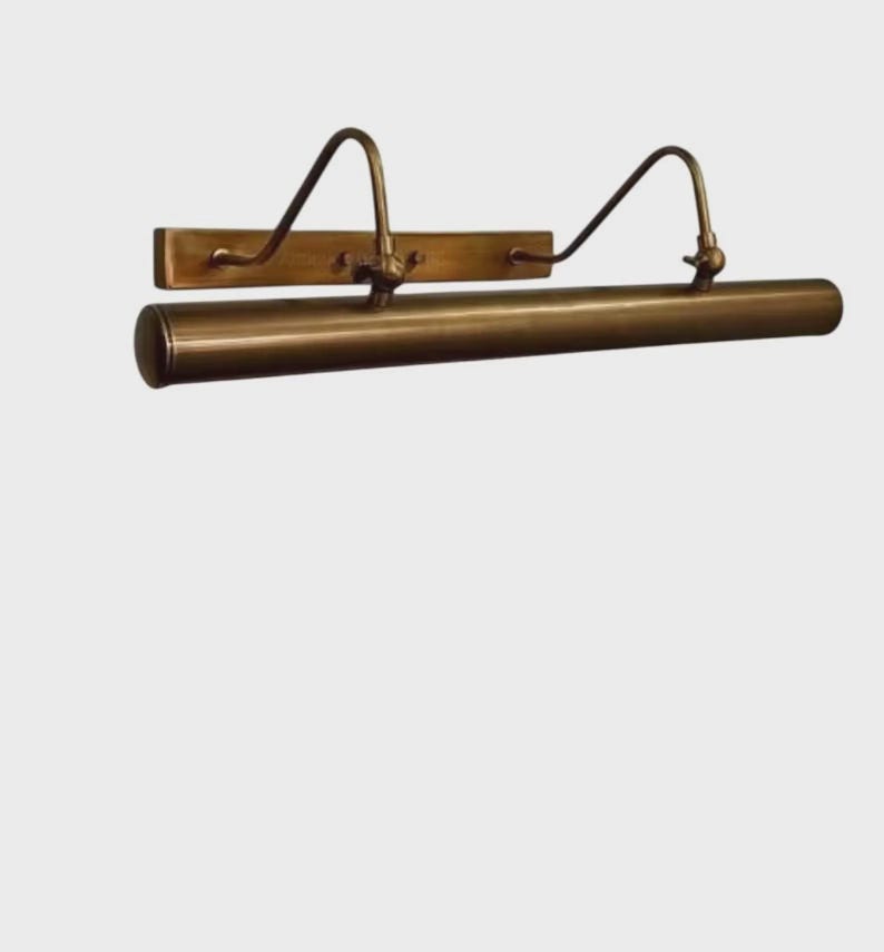 May include: A horizontal picture light fixture with an antique brass finish. The design features a rectangular wall-mounting plate, two curved support arms, and a long cylindrical light housing that extends across the front of the fixture.