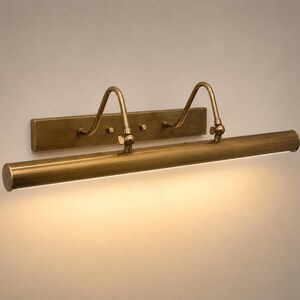 May include: A wall-mounted picture light with an antique brass finish. The fixture features a rectangular backplate, two curved support arms, and a long, cylindrical horizontal tube that emits a warm, downward-facing light across the surface.