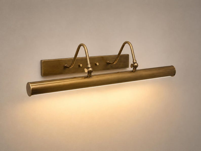 May include: A wall-mounted picture light with an antique brass finish. The fixture features a rectangular backplate, two curved support arms, and a long, cylindrical horizontal tube that emits a warm, downward-facing light across the surface.