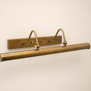 May include: A wall-mounted picture light with a brushed brass finish. The fixture features a long, cylindrical horizontal lamp head attached to a rectangular backplate by two curved, adjustable arms with small wing nuts for positioning.