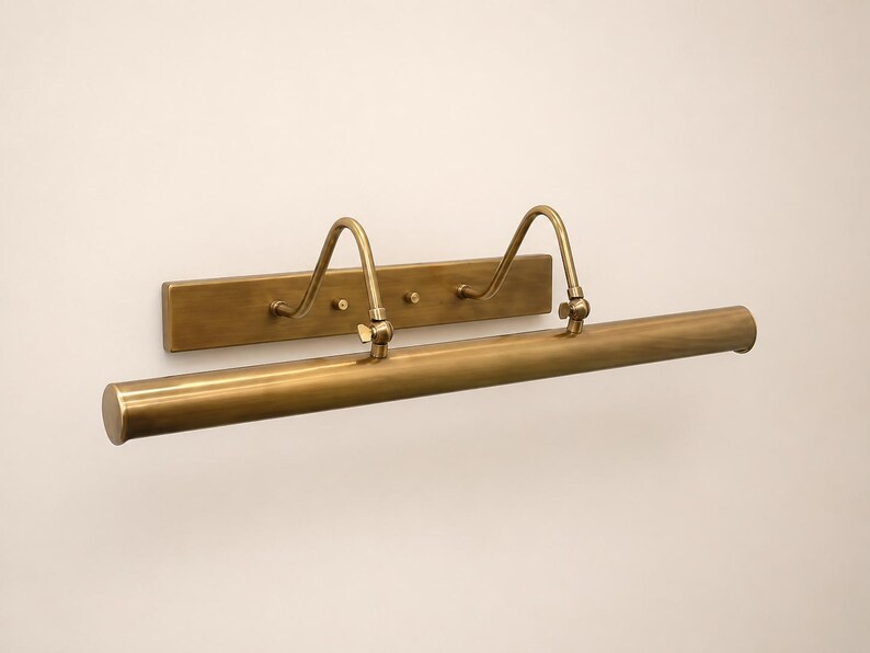 May include: A wall-mounted picture light with a brushed brass finish. The fixture features a long, cylindrical horizontal lamp head attached to a rectangular backplate by two curved, adjustable arms with small wing nuts for positioning.