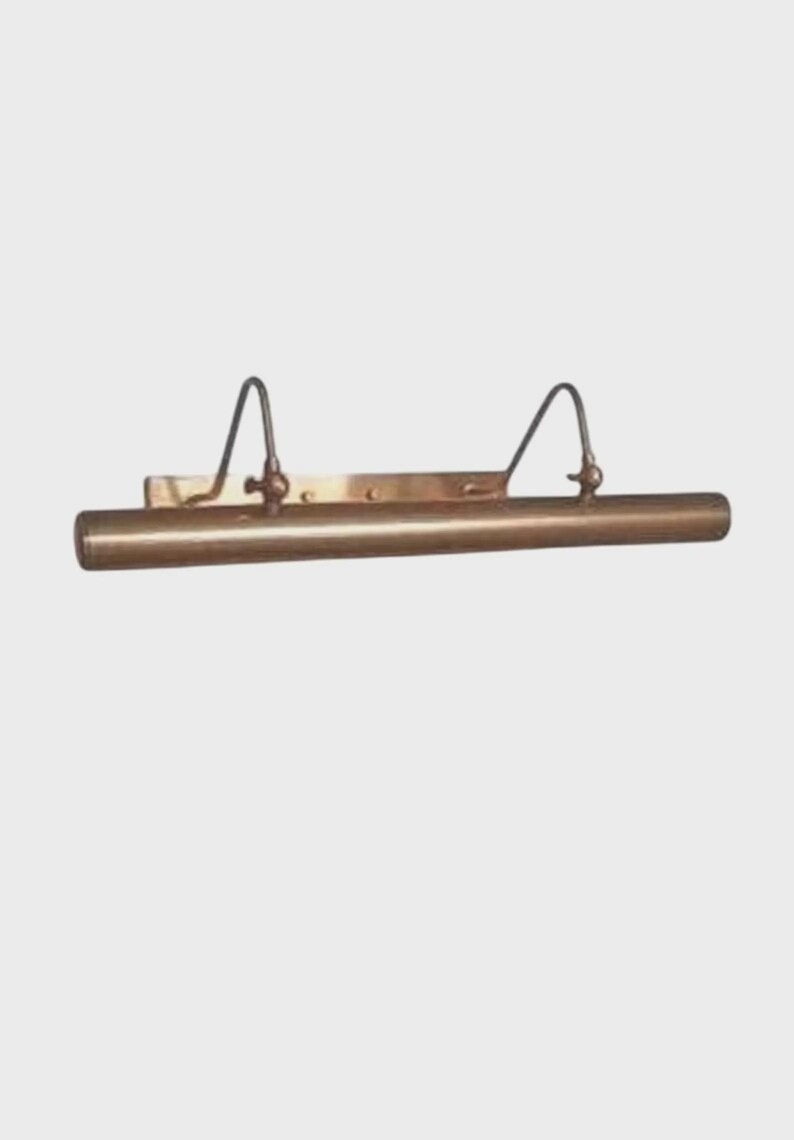 May include: A horizontal, tubular picture light fixture with a metallic bronze finish. The lamp features a rectangular mounting plate and two curved, adjustable arms that connect the base to the cylindrical light housing.