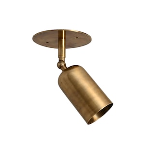 May include: A brass-colored, adjustable spotlight fixture. The fixture has a cylindrical light head and a circular mounting plate. The light head is angled downward, and the fixture is designed for ceiling mounting.