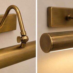 May include: A wall-mounted picture light with a brushed brass finish. The fixture features a rectangular backplate, a curved arm, and a cylindrical light housing with an adjustable wing nut hinge for positioning the angle of the light.