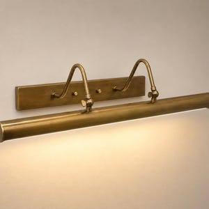 May include: A wall-mounted picture light with an antique brass finish. The fixture features a rectangular backplate, two curved adjustable arms, and a long cylindrical lamp head that emits a warm, downward-facing light across the wall surface.
