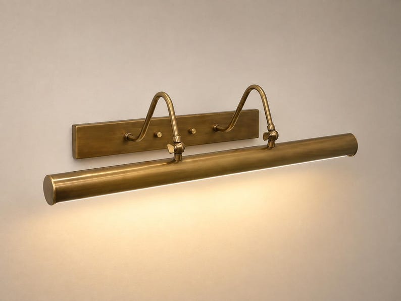 May include: A wall-mounted picture light with an antique brass finish. The fixture features a rectangular backplate, two curved adjustable arms, and a long cylindrical lamp head that emits a warm, downward-facing light across the wall surface.
