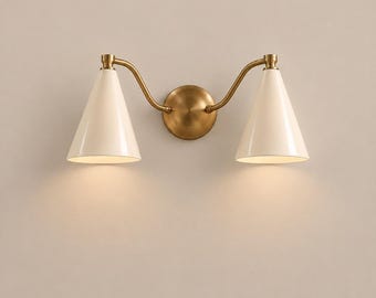 May include: A double wall sconce featuring a central circular brass mounting plate with two curved brass arms extending outward. Each arm supports a downward-facing conical shade in a smooth off-white finish, emitting a soft warm light.