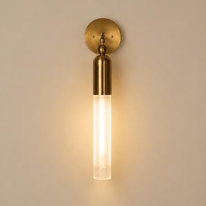 May include: A wall sconce featuring a circular brass backplate and a vertical cylindrical arm. The upper portion of the arm is solid brass, while the lower section consists of a clear, vertically ribbed glass tube emitting a warm light.