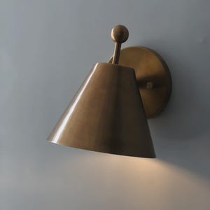 May include: A brass wall sconce with a conical shade and a round backplate. The sconce has a warm, metallic finish and a small, spherical detail at the top. The light shines downward, creating a soft glow.
