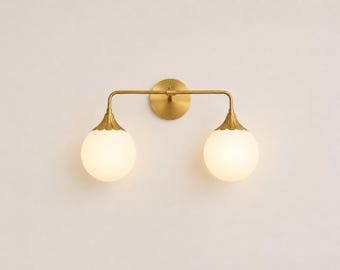 Puede incluir: Un aplique de pared con una barra horizontal de latón unida a una placa circular. Dos brazos orientados hacia abajo se extienden desde la barra, cada uno sosteniendo una pantalla esférica de vidrio blanco con una tapa de latón decorativa.