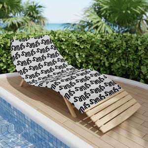 Könnte beinhalten: Ein weißes Strandtuch mit einem sich wiederholenden schwarzen Textmuster, das in fetter serifenloser Schrift "spurs" zeigt. Das Tuch liegt auf einer hölzernen Sonnenliege auf einer Terrasse neben einem Swimmingpool mit blauen Fliesen.