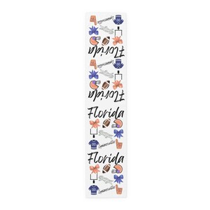 May include: A white table runner featuring a repeating pattern of football-themed illustrations including helmets, footballs, jerseys, goalposts, bows, foam fingers, and the text Florida and Gainesville in a black script font throughout the design.