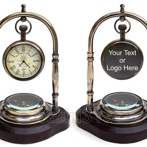 Op de afbeelding: Antieke messingkleurige bureauklok met een kompas en een wijzerplaat met de tekst "Victoria London 1876". De klok is gemonteerd op een donkere houten basis en heeft een decoratief gebogen frame. De wijzerplaat heeft Romeinse cijfers.