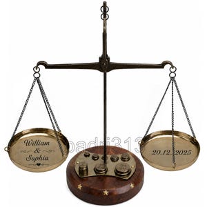 Puede incluir: Una balanza de latón de estilo vintage con base de madera oscura e incrustaciones de estrellas. El plato izquierdo está grabado con "William & Sophia" y un corazón, mientras que el derecho muestra la fecha "20.12.2025". Incluye pesas de latón.