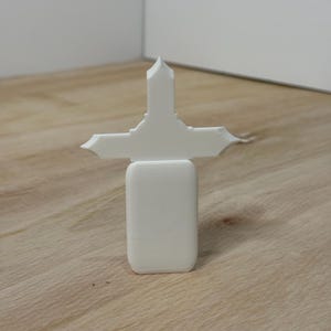 May include: A white, 3D-printed cross-shaped object. The cross has a rectangular base and a detailed top with pointed edges. The object is placed on a light-colored wooden surface.
