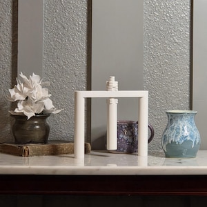 May include: A white, square-framed object with a central, vertical post and a stack of small, white, cylindrical pieces on top. A ceramic vase with white flowers, a mug, and a blue and white vase are also visible.