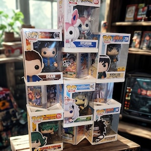 May include: A stack of Funko Pop! figures in their original boxes. The figures are from various franchises, including Pokémon, Supernatural, Naruto, and Genshin Impact. The boxes are clear plastic, and the figures are visible inside. The figures are displayed on a wooden surface.