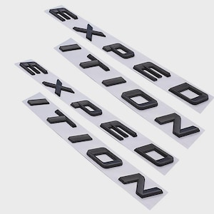 Op de afbeelding: Vier sets zwarte plastic letters die "EXPEDITION" spellen op witte plakstrips. De letters zijn dik en driedimensionaal, met een matte afwerking. De achtergrond is effen lichtgrijs.