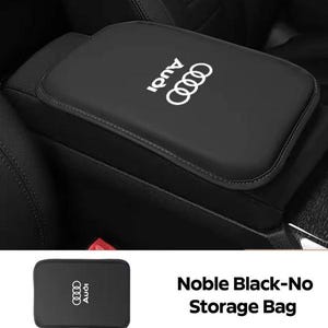 May include: Black car armrest cover featuring the Audi logo in white. The cover is rectangular and designed to fit over a car's centre console armrest. A small storage bag with the Audi logo is also visible.