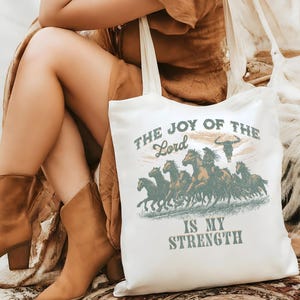 May include: White tote bag with the text "THE JOY OF THE LORD IS MY STRENGTH" and a graphic of running horses. The design is in shades of brown and green. The bag has long white handles.