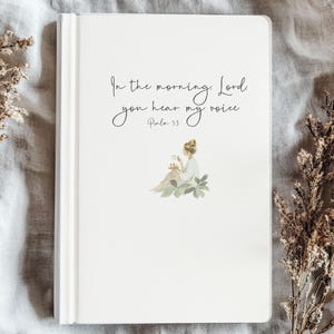 May include: A white journal with the quote "In the morning, Lord, you hear my voice" and Psalm 5:3 printed on the cover. The cover features a watercolour illustration of a woman with flowers. The journal is closed and placed on a textured surface.