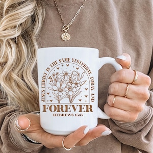Może przedstawiać: Biały ceramiczny kubek z uchwytem. Kubek ma napis "JESUS CHRIST IS THE SAME YESTERDAY, TODAY, AND FOREVER HEBREWS 13:8" w kolorze brązowym, wraz z kwiatowym wzorem. Kubek jest trzymany przez osobę.