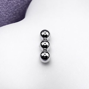 May include: Three small, shiny, silver ball earrings arranged vertically. The earrings are set against a white, cloud-shaped background, with a soft, purple fabric in the upper left corner. The earrings are likely for pierced ears.