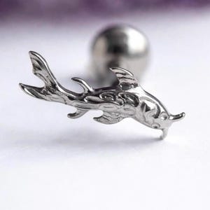 May include: A silver-toned metal stud earring shaped like a fish with detailed fins and a tail. The earring features a smooth, reflective surface and is attached to a small, round metal ball backing, creating a distinct aquatic-themed jewellery piece.