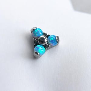 May include: A silver-toned, triangular body jewellery piece featuring three blue opal spheres and a central silver sphere. The piece is designed with a geometric shape and is suitable for body piercings.