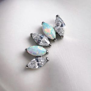 May include: Close-up of several pieces of jewellery. Each piece features a marquise-cut gemstone, alternating between clear, faceted stones and opalescent stones. The settings are silver-toned, and the pieces are arranged on a white surface.