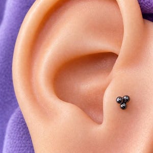 May include: Close-up of an ear with a small, dark stud earring. The earring has a cluster of three small, round beads. The skin tone is light, and the background is a soft purple.