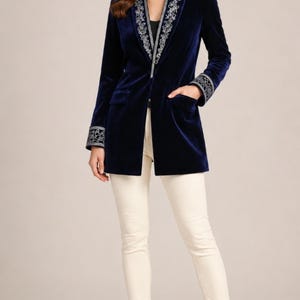 May include: A deep navy blue velvet blazer featuring a shawl collar and cuffs adorned with intricate silver floral embroidery. The blazer has a long-line silhouette, two front flap pockets, and is paired with slim-fit cream trousers and black heels.